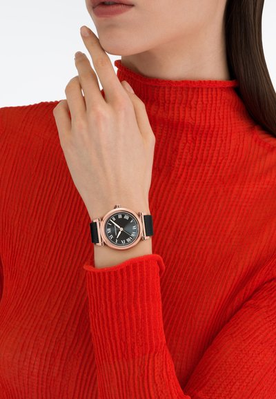 Ženska v rdeči rebrasti majici se dotika brade, na levi zapestju nosi uro iz rožnatega zlata s črnim usnjenim paščkom in črnim številčnico z rimskimi številkami.