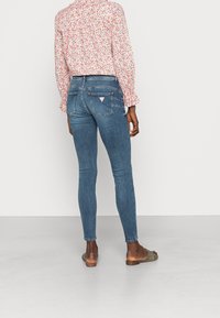 Jeans de denim azul ajustados con una cintura de altura media, que presentan bolsillos traseros y un pequeño parche con el logo. El modelo lleva una blusa floral de manga larga.