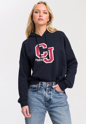 Granatowa bluza z kapturem z dużym czerwono-białym logo "CJ". Posiada kaptur ze sznurkiem i ściągacze przy rękawach, zestawiona z jasnoniebieskimi dżinsami.