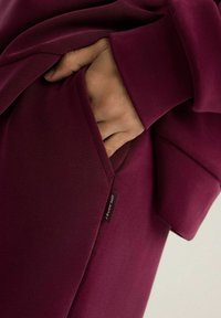 Bluza z długim rękawem w głębokim burgundowym kolorze z kieszenią z przodu. Miękka tkanina, luźny krój, z metką na dolnym brzegu. Gładka, elastyczna faktura.