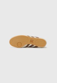 Brun og hvid sneakersål med tekstureret gummi, der har et cirkulært mønster og tre vandrette striber. Adidas-logo prægede.