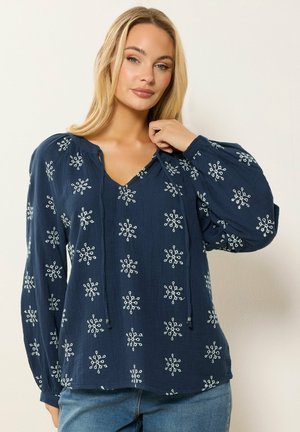 Frau trägt eine marineblaue Bluse mit weißen, bestickten Schneeflockenmuster und Ballonärmeln, kombiniert mit blauen Jeans, vor einem schlichten Hintergrund.