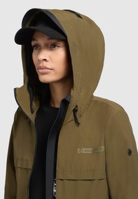 Giacca con cappuccio verde oliva con chiusura a zip, dotata di un patch del logo nero e coulisse regolabili sia sul cappuccio che sull'orlo.