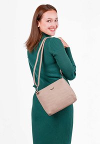 Béžová kabelka přes rameno s texturovaným prošívaným vzorem, nastavitelným popruhem a jemným detailem loga. Modelka má na sobě zelené žebrované šaty.