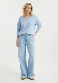 Hellblaues V-Ausschnitt-Pullover aus weichem Material, kombiniert mit hochgeschnittenen, hellblauen weit geschnittenen Jeans. Das Model trägt graue Sneaker.