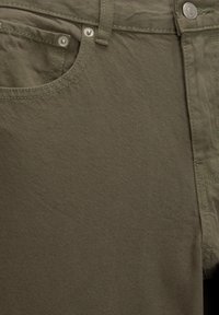 Pantalon en mélange de coton vert olive avec une finition texturée, présentant des coutures apparentes, une poche avant et des accents de boutons métalliques.