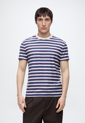 Junger Mann trägt ein kurzärmeliges weißes T-Shirt mit blauen und roten horizontalen Streifen und dunkle Hose, steht vor einem schlichten Hintergrund.