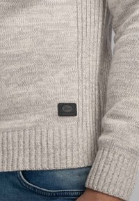 Maglione grigio chiaro lavorato a maglia con texture a coste. Presenta una toppa con logo nera e un orlo aderente. Abbinato a pantaloni jeans blu.