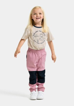Lachend kind met lang blond haar dat een beige t-shirt draagt met outdoorprint, roze en zwarte broek en witte sneakers, staand met licht gespreide armen.