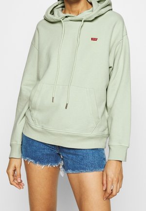Personne portant un sweat à capuche Levi's vert clair avec poche avant et cordons, associé à un short en jean bleu effiloché.