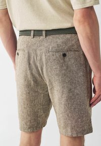 Shorts de lino marrón con una tela texturizada de color marrón claro. Presenta un cinturón tejido en negro y verde y dos bolsillos traseros con botones.