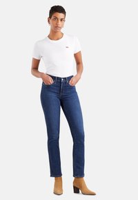 White short-sleeve t-shirt with red logo, paired with dark blue high-waisted jeans and tan heeled ankle boots. Simple, fitted design.