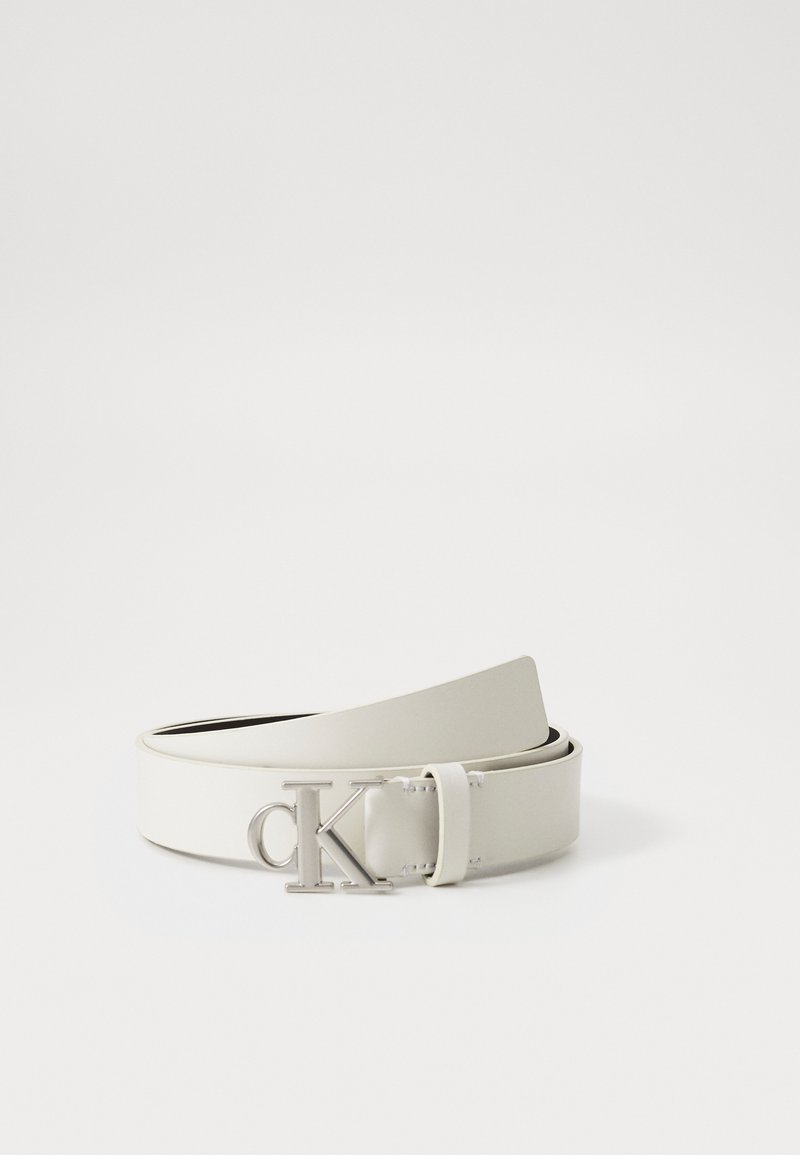 Fehér bőr öv sima textúrával, ezüst színű csattal, amelyen a "CK" monogram látható. Állítható hosszúságú, modern dizájnnal.