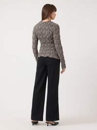 Femme se tenant de profil, portant un haut taupe à manches longues texturé, un pantalon large noir et des chaussures à talons noires, sur fond blanc.