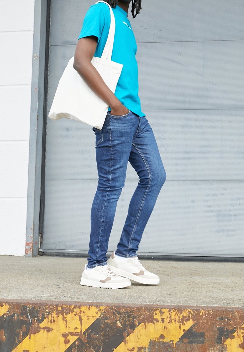 Camiseta de algodón azul, bolso de lona blanco, jeans ajustados y zapatillas blancas con detalles beige sobre un fondo gris.