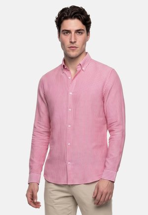Uomo che indossa una camicia a maniche lunghe a righe verticali rosa e bianche con pantaloni beige, in piedi davanti a uno sfondo semplice.
