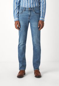 Jeans in denim azzurro chiaro con una vestibilità dritta, tasche frontali e cuciture classiche. Indossati con scarpe in pelle marrone e abbinati a una camicia a righe.