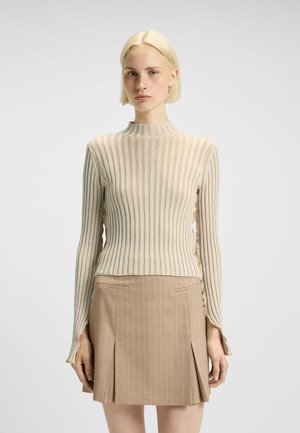 Lichtbeige gebreide trui met lange mouwen en hoge hals, gecombineerd met een lichtbruine minirok met streepjes en zijzakken.