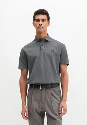 Mees kannab tumerohelist lühikese varrukaga polomeest, mis on tumesinine ja sisenõelaga hallidesse voltpükstesse pistetud, musta vööga, valgel taustal.