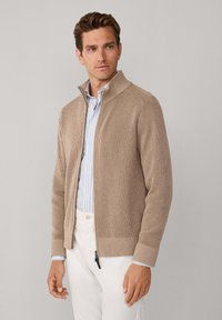 Beige geribbelde gebreide jas met een hoge kraag, ritssluiting aan de voorkant en lange mouwen, gedragen over een lichtblauw gestreept overhemd en een witte broek.