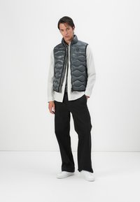 Gray puffer vest with quilted pattern, front zipper, and ribbed collar, worn over a white button-up shirt and black pants. White sneakers.
