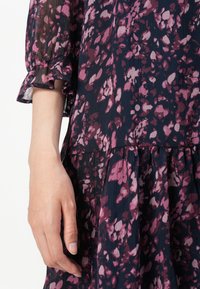 Blomstrete kjole i mørk marineblå med lilla og rosa detaljer. Lett stoff med plisserte teksturer og volanger ved ermeåpningene.