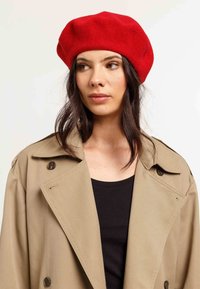 Beret roșu din lână cu o textură moale, formă rotunjită, purtat cu un trench coat bej și un top negru. Prezintă un design clasic și simplu.