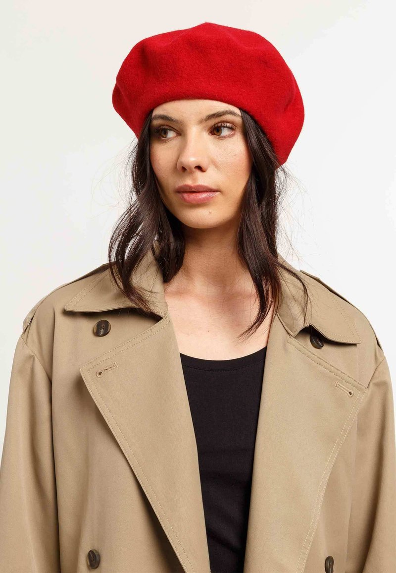 Beret roșu din lână cu o textură moale, formă rotunjită, purtat cu un trench coat bej și un top negru. Prezintă un design clasic și simplu.