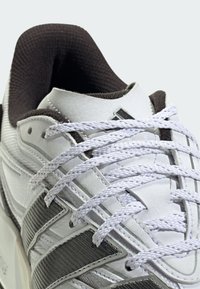 White sneaker with textured synthetic material, featuring gray accents, white laces, and a padded collar. Prominent stitching details visible.