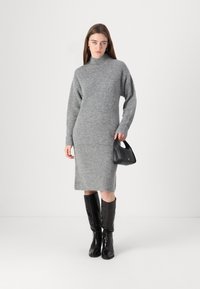 Gestricktes graues Kleid mit hohem Kragen und langen Ärmeln, das bis zur Mitte der Waden reicht. Kombiniert mit kniehohen schwarzen Stiefeln und einer kleinen schwarzen Handtasche.