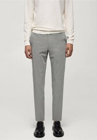 Homme portant un pantalon slim gris clair, des chaussures noires à lacets et un pull à manches longues écru, debout devant un fond uni.