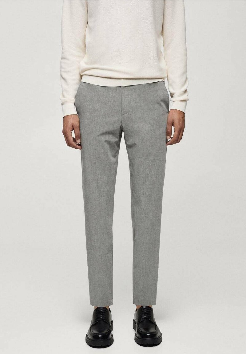 Homme portant un pantalon slim gris clair, des chaussures noires à lacets et un pull à manches longues écru, debout devant un fond uni.