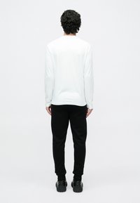 Άνδρας με σγουρά μαλλιά που φοράει λευκό πουκάμισο με μακριά μανίκια, μαύρο παντελόνι και μαύρα παπούτσια, στέκεται με την πλάτη γυρισμένη σε απλό φόντο.