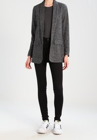 Grå texturerad blazer med öppen framsida, två fickor, kombinerad med en svart skjorta och smala svarta byxor. Skorna är vita sneakers med svarta detaljer.