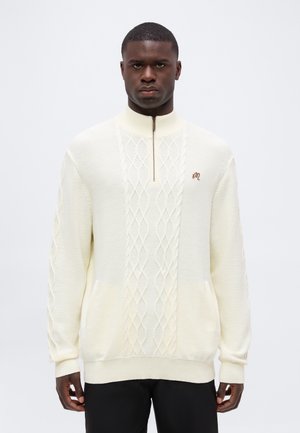Homme portant un pull crème en maille torsadée avec un col zippé et un petit logo brodé sur la poitrine, debout devant un fond blanc.