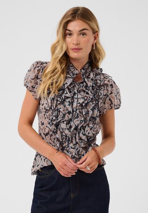 Ung kvinde med langt blondt hår iført en mønstret bluse med flæser og mørke jeans, stående med hænderne foldet foran.