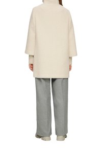 Beige, gestructureerde oversized jas met een hoge kraag, dropped shoulders en wijde mouwen; gecombineerd met grijze op maat gemaakte broek en beige schoenen.