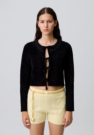 Cardigan nero corto realizzato in un tessuto morbido, con scollatura tonda e chiusure a lacci al centro. Abbinato a pantaloni corti in maglia color crema.