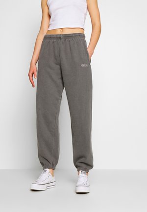 Person trägt graue Sweatpants mit elastischem Bund und BDG-Logo, weiße Plateau-Sneaker und ein weißes kurzes Tanktop, steht vor einem schlichten Hintergrund.