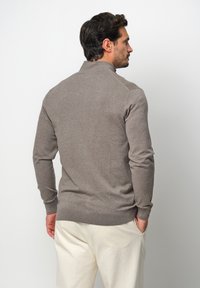 Maglione marrone con colletto alto, maniche lunghe, polsini e orlo ribattuti. Texture morbida, design minimalista. Indossato con pantaloni chiari.