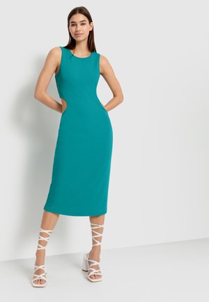Ein tailliertes, ärmelloses türkisfarbenes Kleid mit seitlichen Ausschnitten und schicker Midi-Länge, kombiniert mit weißen, schnörkeligen Blockabsätzen. Glatte Stofftextur.