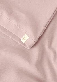 Jasnoróżowa bawełniana tkanina o miękkiej, gładkiej teksturze, z małą kremową metką w rogu. Tkanina jest ułożona w fałdy, ukazując zagięcia.