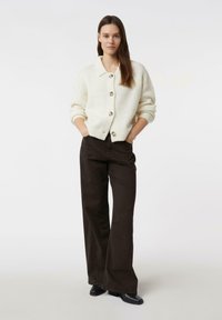 Ecru sweter-kardigan z grubymi guzikami, zestawiony z ciemnobrązowymi rozszerzanymi spodniami. Model stoi z rękami w kieszeniach na jednolitym tle.