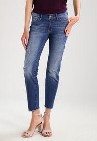 Denimjeans i medelblått, med slitna lappar, slim fit, medellågt midjeband och fransad fåll. Matchas med sandaler med blockklack i ljusgrått.