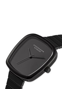 Sort firkantet ur med en glat mat overflade; urskiven viser minimalistiske time- og minutvisere og har mærkenavnet "Copenhagen Studios."