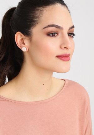 Roséguld studs örhängen med en slät, rund design. Bärs av en modell med en mjuk rosa topp och rakt, mörkt hår i en hästsvans.