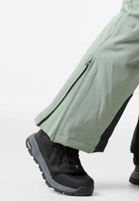 Lichtgroene waterdichte broek met zijritsen en zwarte accenten, gecombineerd met zwarte wandelschoenen met robuuste zolen en gestructureerde details.