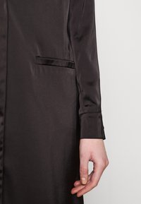 Bras droit d’une personne portant un vêtement long en satin noir avec une petite poche fendue sur le côté, sur un fond clair.