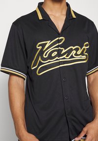 Musta lyhythihainen napitettava paita, jossa on keltainen kaulus, valkoiset raidat hihoissa ja kultalangalla brodeerattu "Kani" -teksti etuosassa.