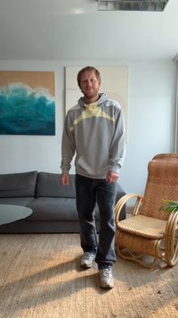 Hoodie cinza claro com detalhes amarelos, jeans escuros e ténis cinzas. Num interior moderno com um sofá cinza e uma cadeira de vime.
